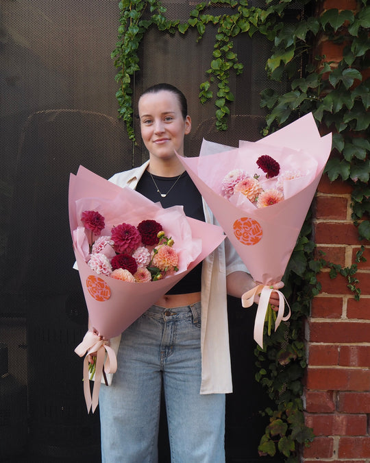 Person holding two bouquets of flowers against a brick wall with greenery.
