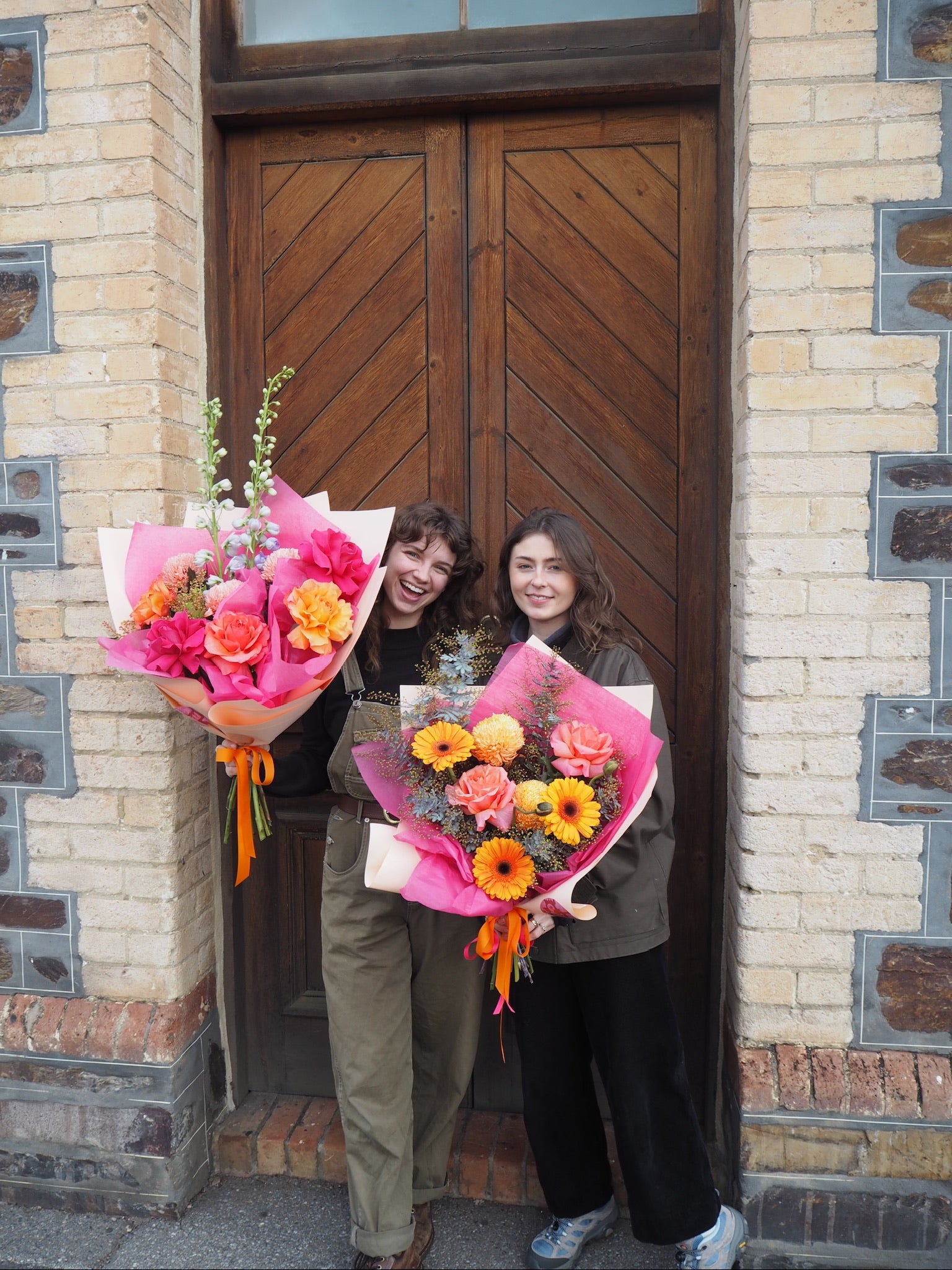 Two people holding flower bouquets standing in front of a wooden door.