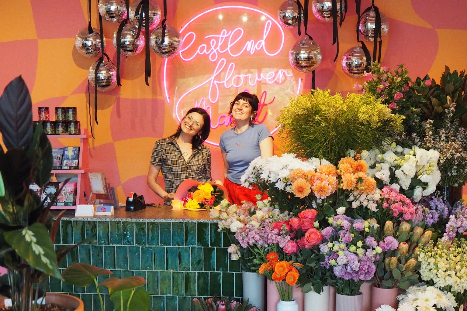 Two people behind a floral counter with colorful flowers and a neon sign in the background.