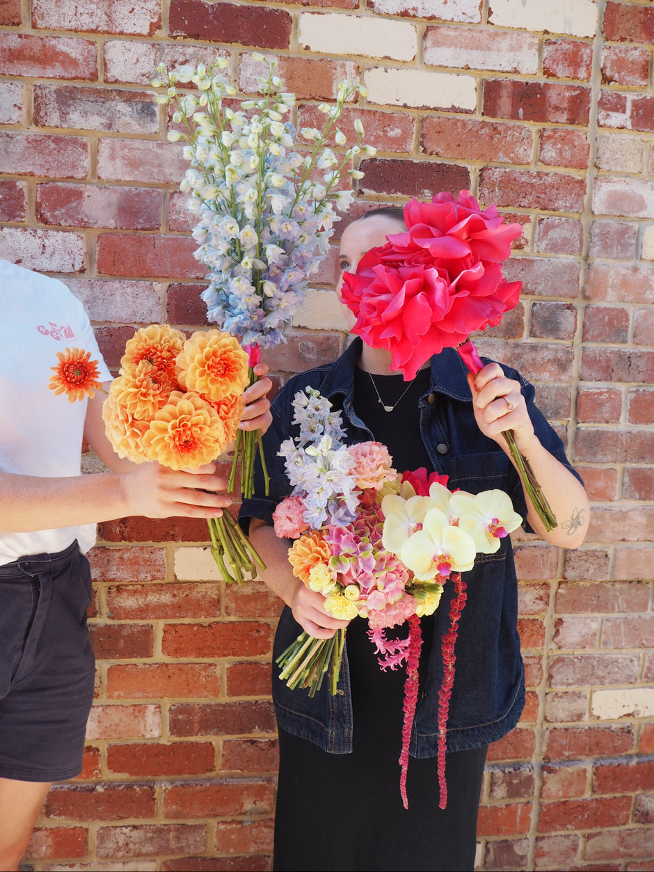 Two people holding colourful bridal flower bouquets against a brick wall.