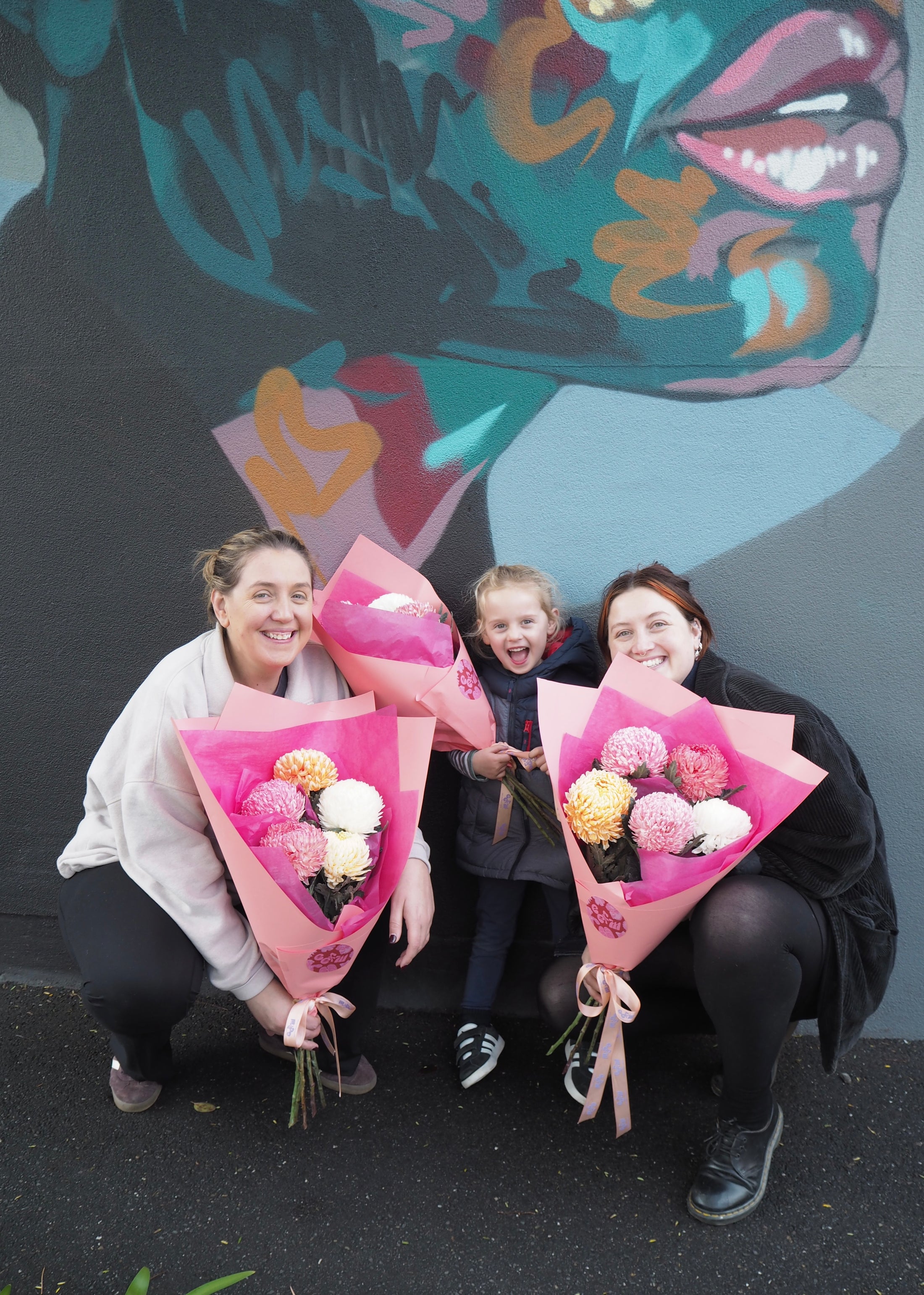 Three people holding chrysanthemum bouquets in front of a colourful mural.