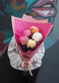 Load image into Gallery viewer, Person holding a bouquet of flowers wrapped in pink paper against a colorful background
