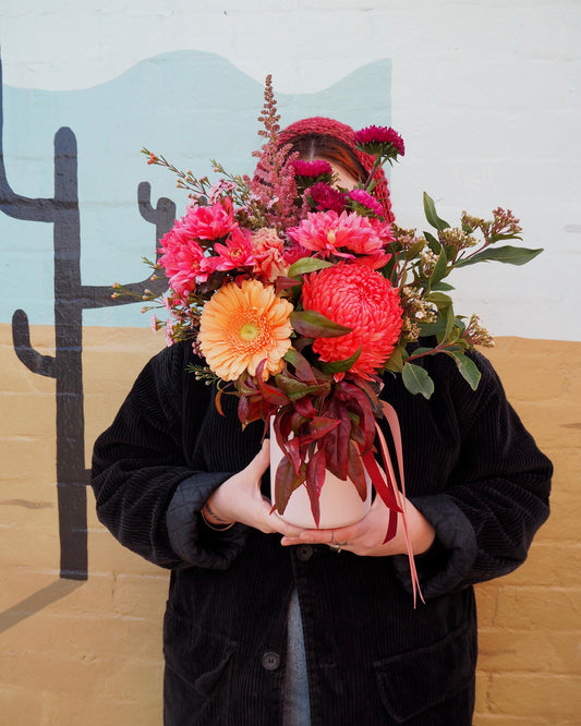 Person holding a ceramic vase filled with flowers in orange, hot pink and coral tones.