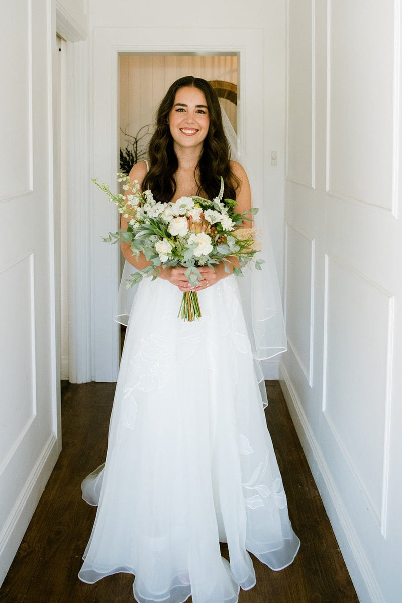 Woman in a white dress holding a bouquet in a hallway with a mirror reflection.