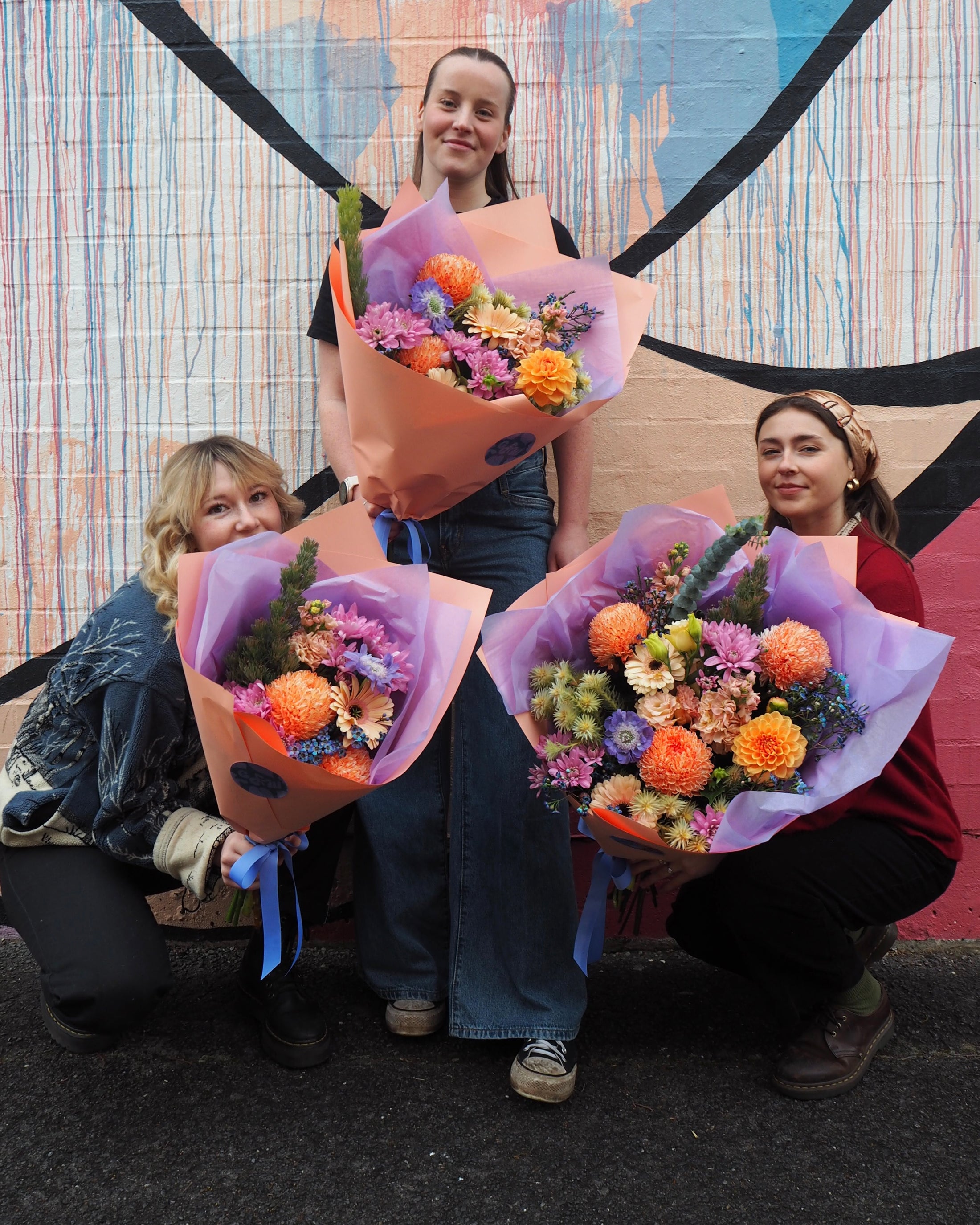 Three people holding colorful flower bouquets in front of a vibrant mural.