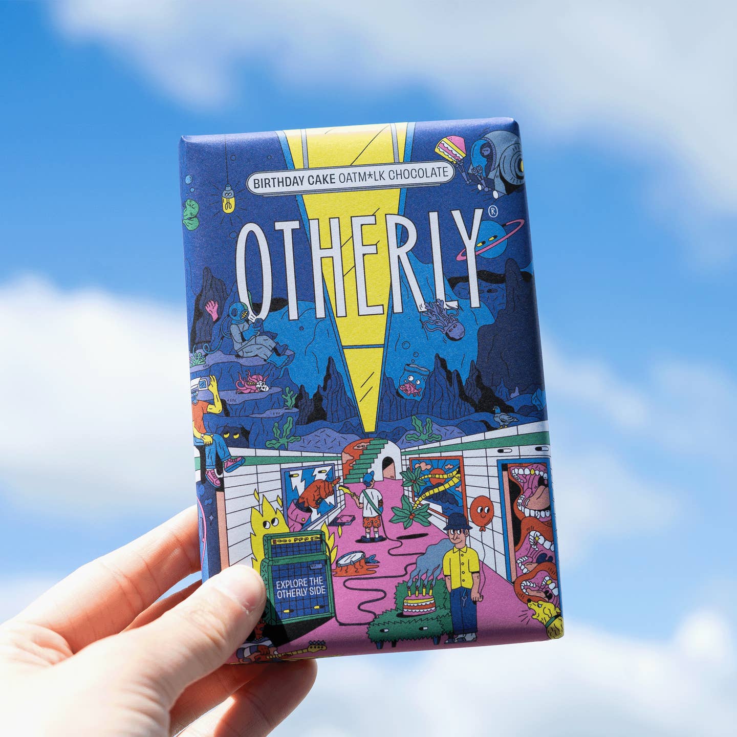 A hand holding a bar of OTHERLY Vegan Chocolate with a colorful and illustrated packaging depicting various characters and a birthday cake.