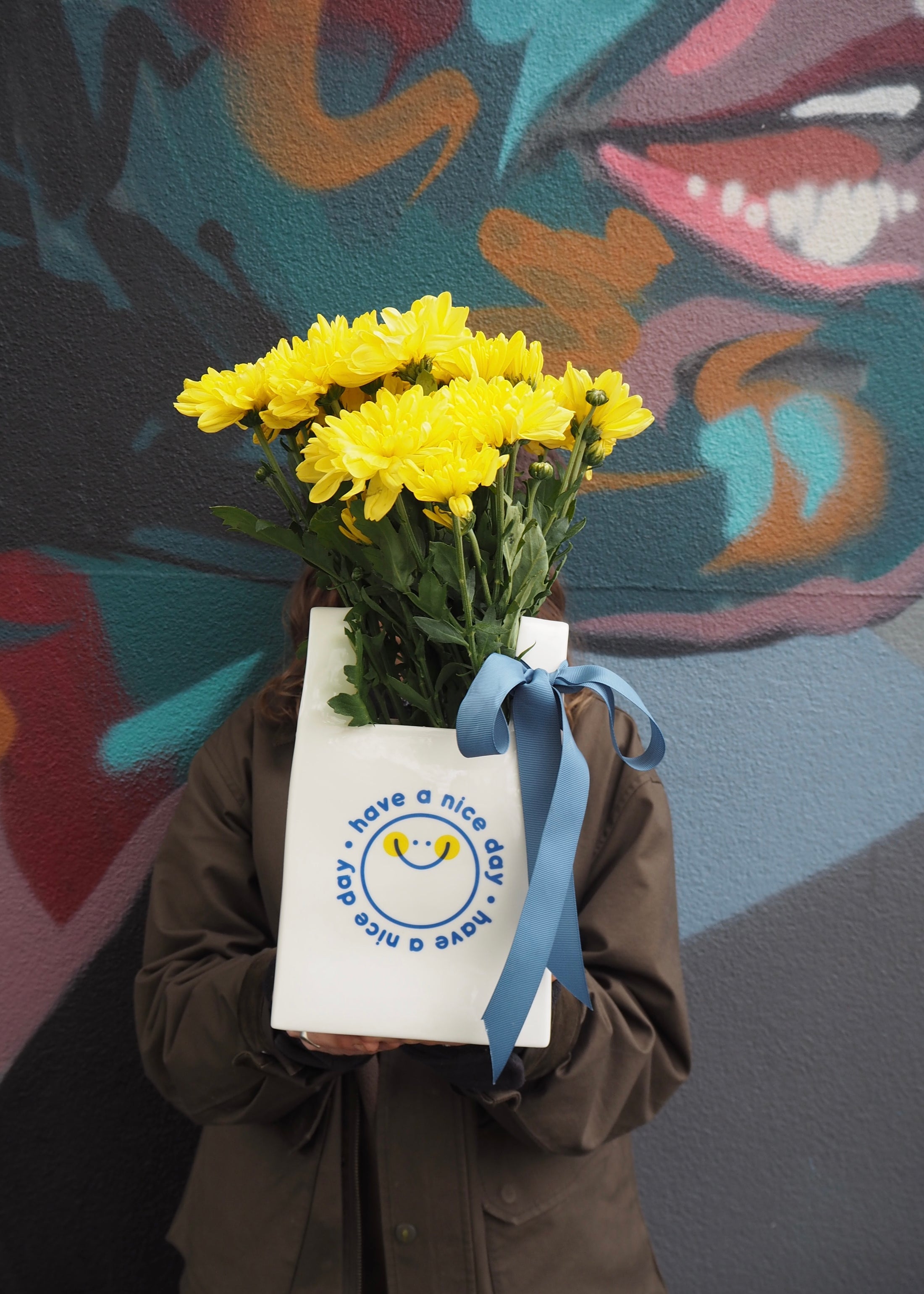 Person holding a bouquet of yellow flowers wrapped in white paper with a blue ribbon against a colorful mural.