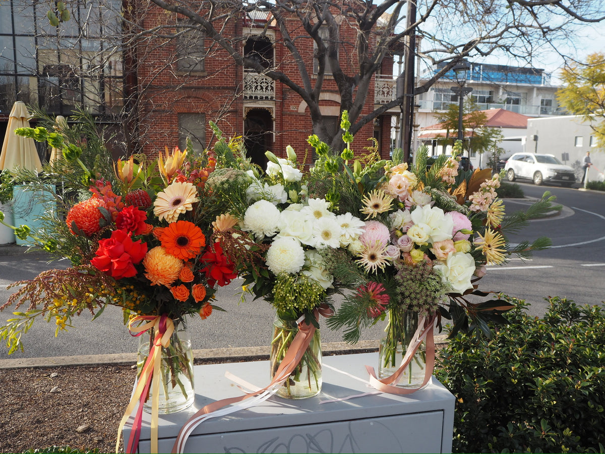 Three floral arrangements on a table with a building and street in the background